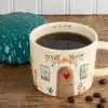 Colorful ceramic mug with house and floral design filled with black coffee, placed on a wooden table with coffee beans.