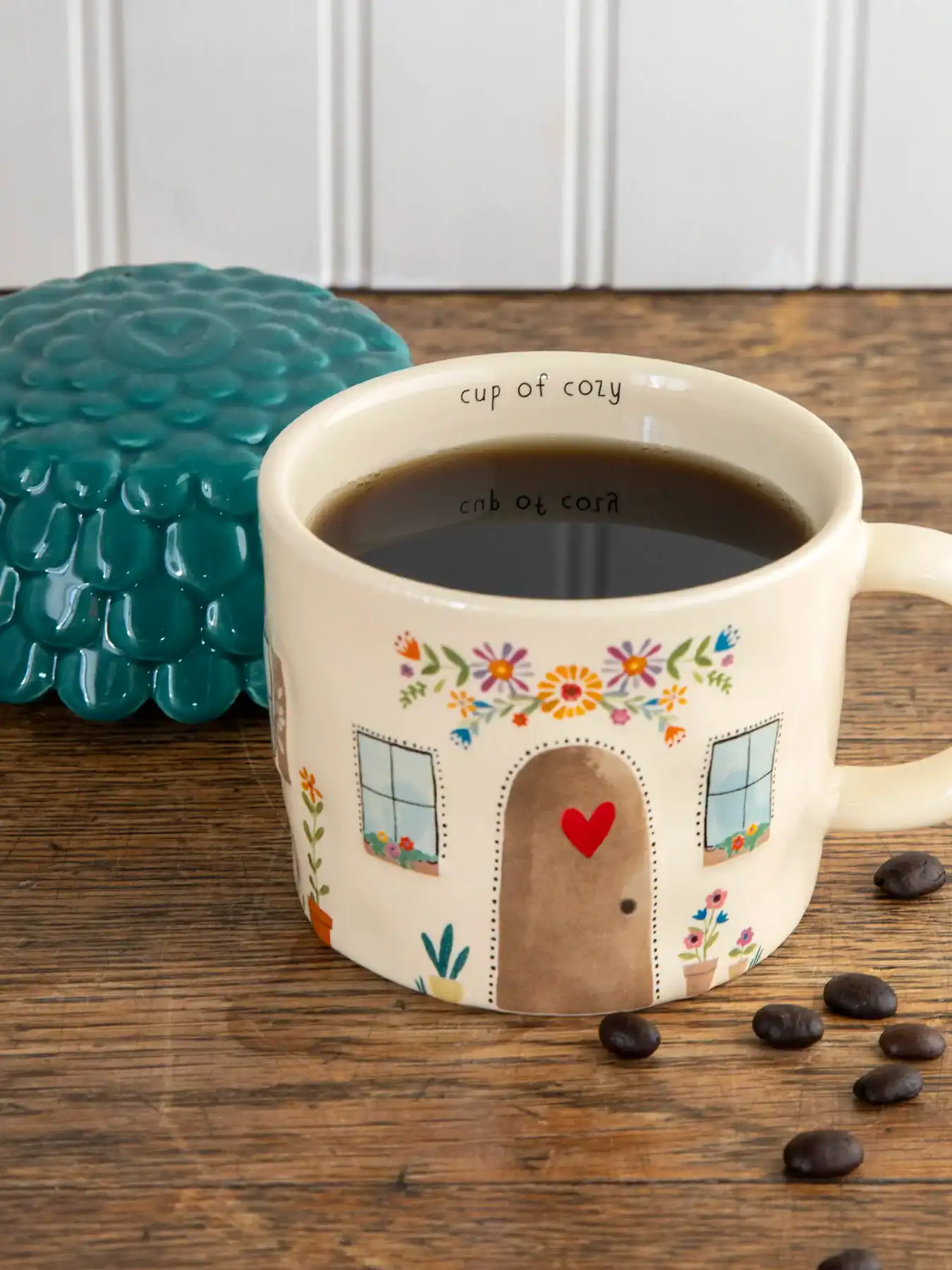 Colorful ceramic mug with house and floral design filled with black coffee, placed on a wooden table with coffee beans.