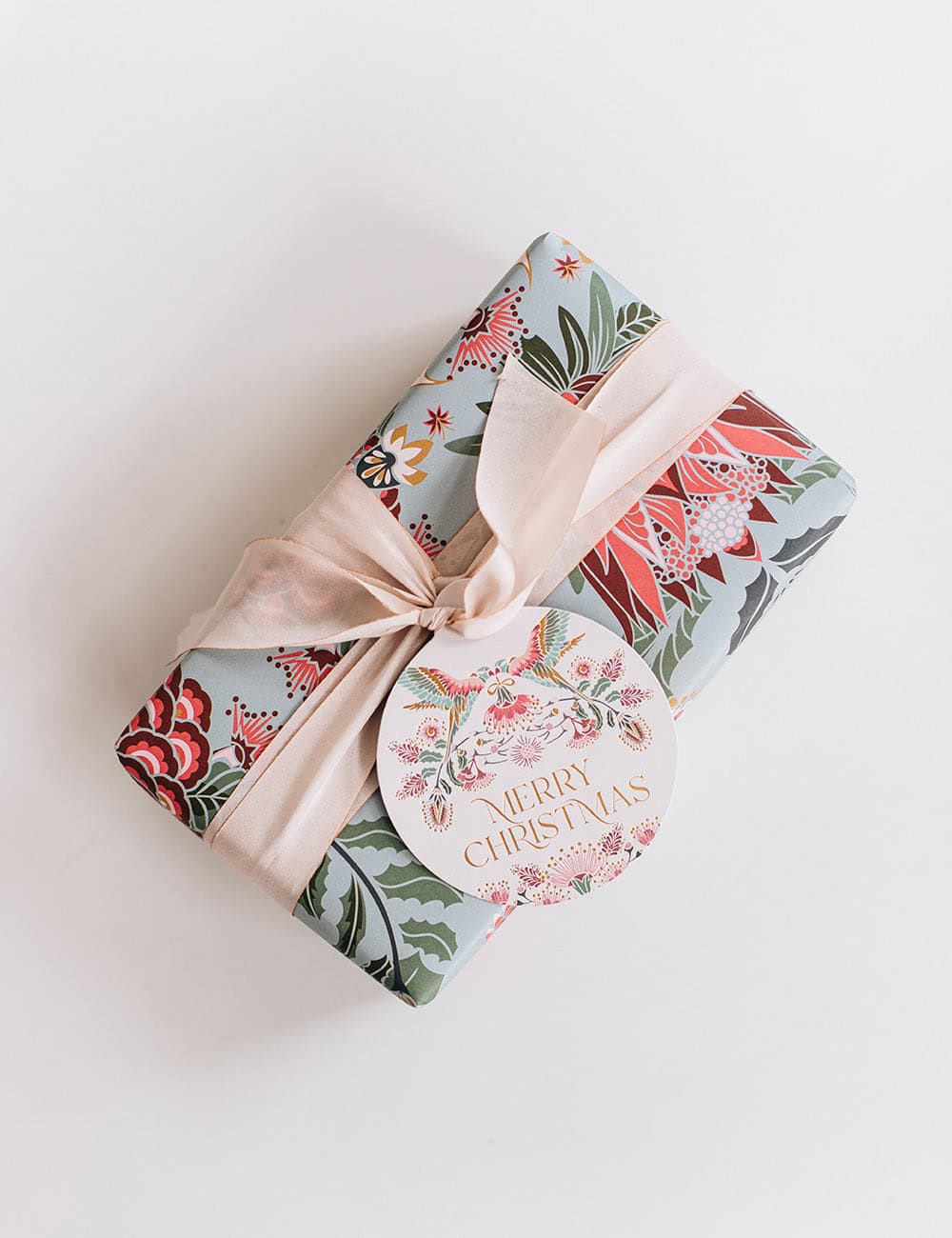 Festive gift wrapped with holiday-themed floral paper, pale pink satin ribbon, and a "Merry Christmas" round gift tag, perfect for Christmas presents from The Wraperie.