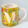 Colorful floral mug with a large white letter "M" and the phrase "cup of happy" inside, perfect for cheerful mornings and personalized gift-giving.