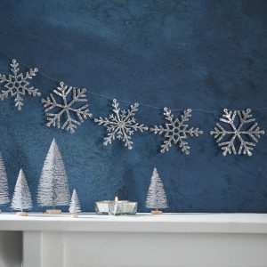 Silver glitter snowflake garland hanging on a dark blue textured wall, holiday decoration with small white felt Christmas trees and a lit candle on a white shelf, festive winter decor.