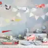 Colorful paper airplane and cloud-themed party decorations for kids' birthday celebration, featuring a sky backdrop, bunting flags, and themed tableware. Perfect for aviation-themed parties.