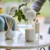 White candle in a modern ceramic holder on a marble table with white flowers and diffuser, elegant home decor for relaxation and ambiance at The Wraperie.
