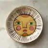 Vibrant decorative ceramic plate with artistic face design, featuring bold colors and gold accents, perfect for stylish home decor or gifting at The Wraperie.