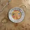 Colorful decorative plate with a face design on a wooden surface, surrounded by jewelry pieces. Perfect for home decor, wall art, and unique accent pieces from The Wraperie.