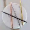 Polished metal pens in gold, silver, rose gold, and black colors arranged on a round white marble surface. Perfect promotional stationery for The Wraperie, combining elegance and branding.