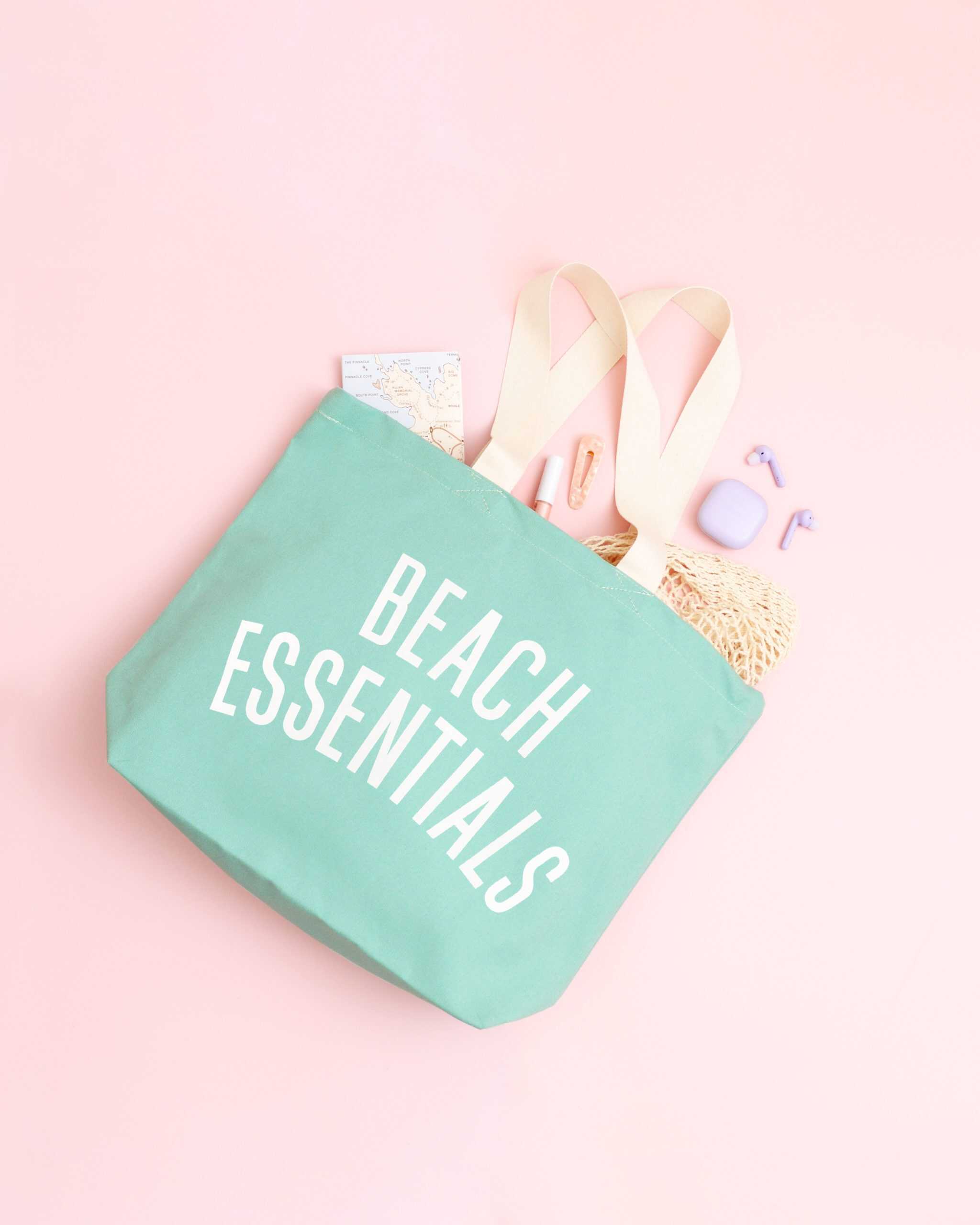 Reusable beach tote bag with "Beach Essentials" printed on it, filled with summer accessories and items, on a pastel pink background.