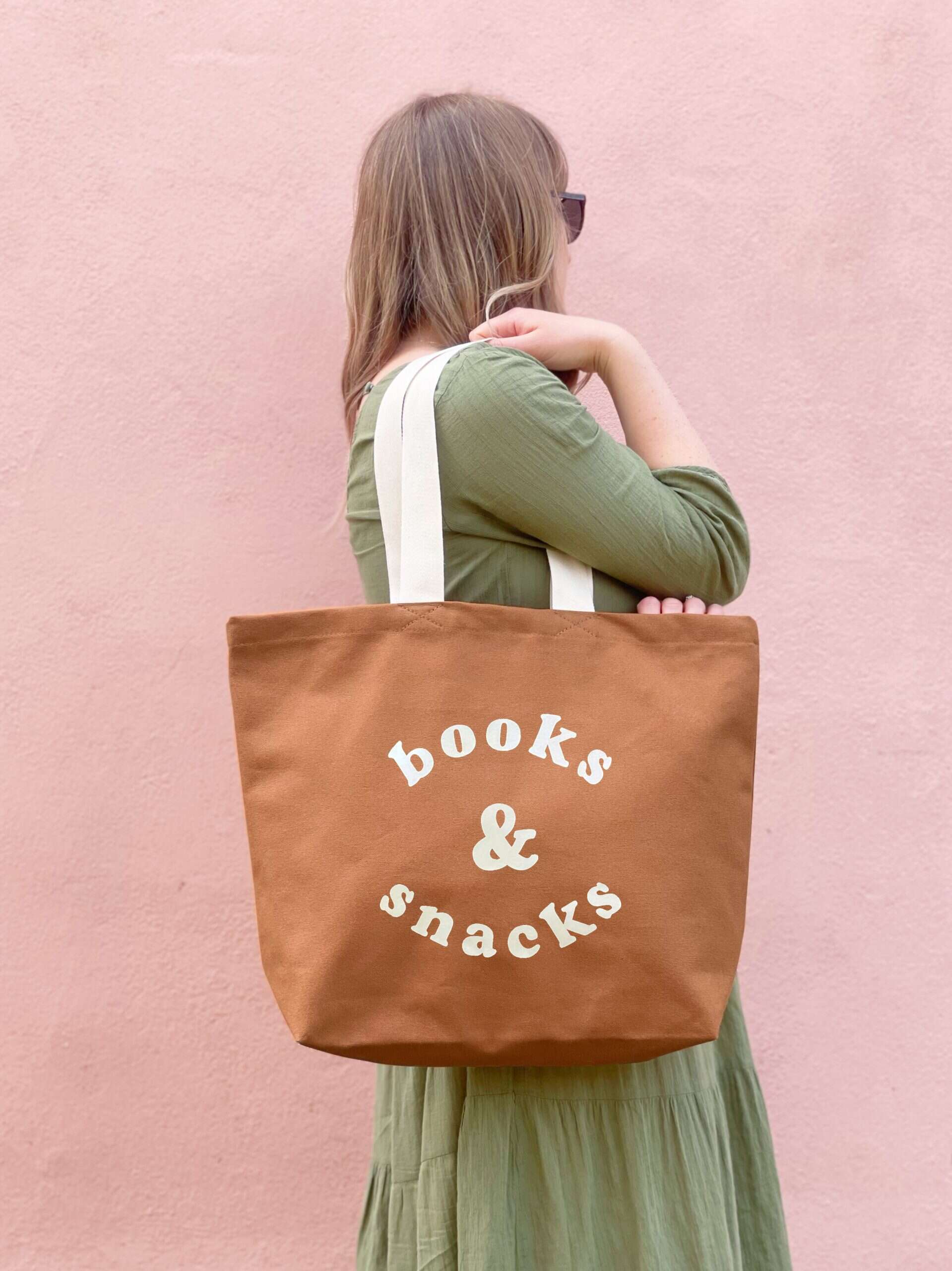Eco-friendly tote bag with "Books & Snacks" text, perfect for reading enthusiasts and snack lovers, on a woman against a pink background.