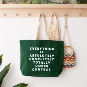 Vibrant green tote bag with bold white text reading "Everything is absolutely completely totally under control" hanging on a wall hook with other stylish bags, in a bright, modern art and home decor shop.