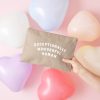 Bright pink and pastel heart-shaped balloons around a beige clutch bag with "Exceptionally Wonderful Human" printed on it, promoting positivity and celebration for special occasions.