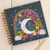 Vivid notebook featuring a colorful rainbow, moon, stars, and floral design, perfect for journaling or note-taking at The Wraperie.