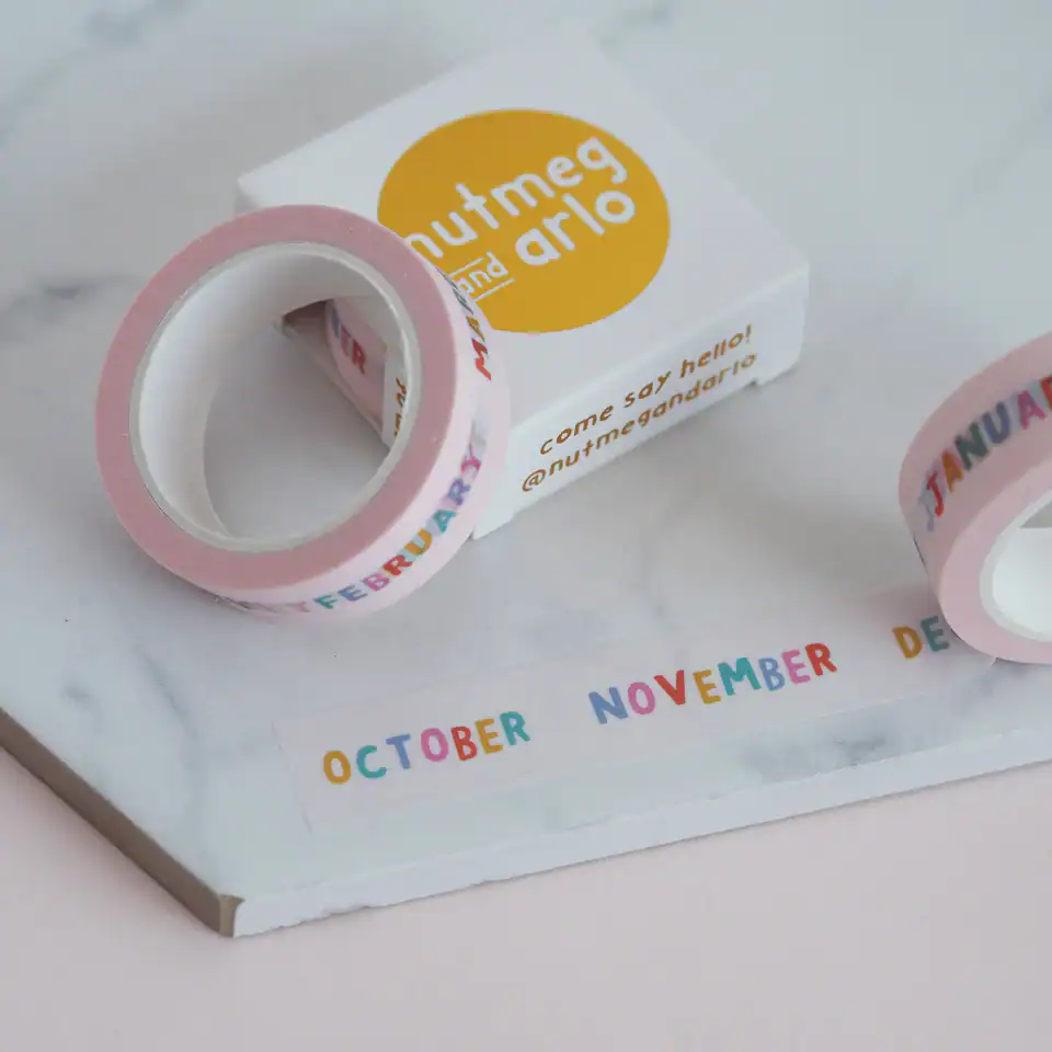 Colorful washi tape with months of the year printed in rainbow colors, displayed alongside a small box with the "Nutmeg and Arlo" logo.