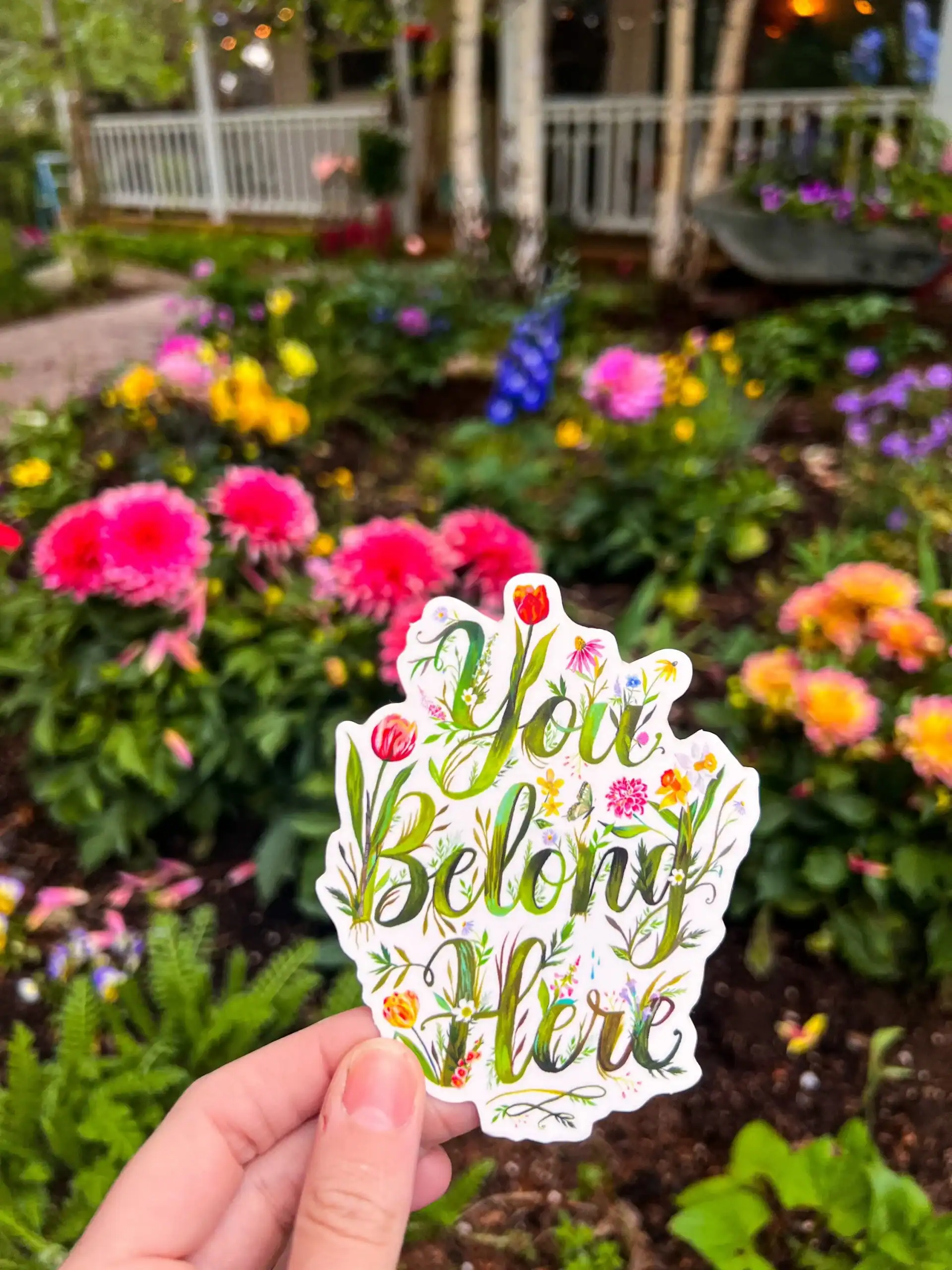 Colorful flower garden with a "You Belong Here" sticker held in hand, showcasing a welcoming atmosphere at The Wraperie. Beautiful blooms and vibrant garden setting emphasize freshness and community in the space.